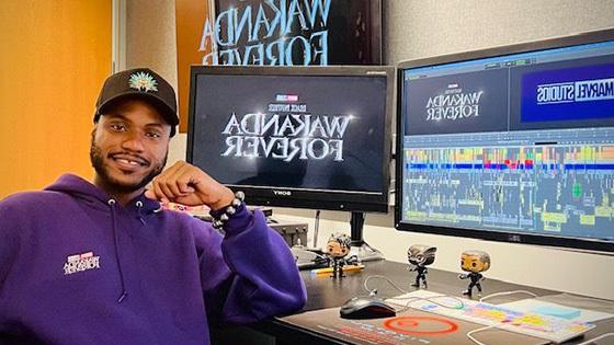 阿兰坐在一张桌子前，桌子上有多个显示器，上面写着“永远的瓦坎达”和“漫威工作室”.” He is wearing a purple hoodie.